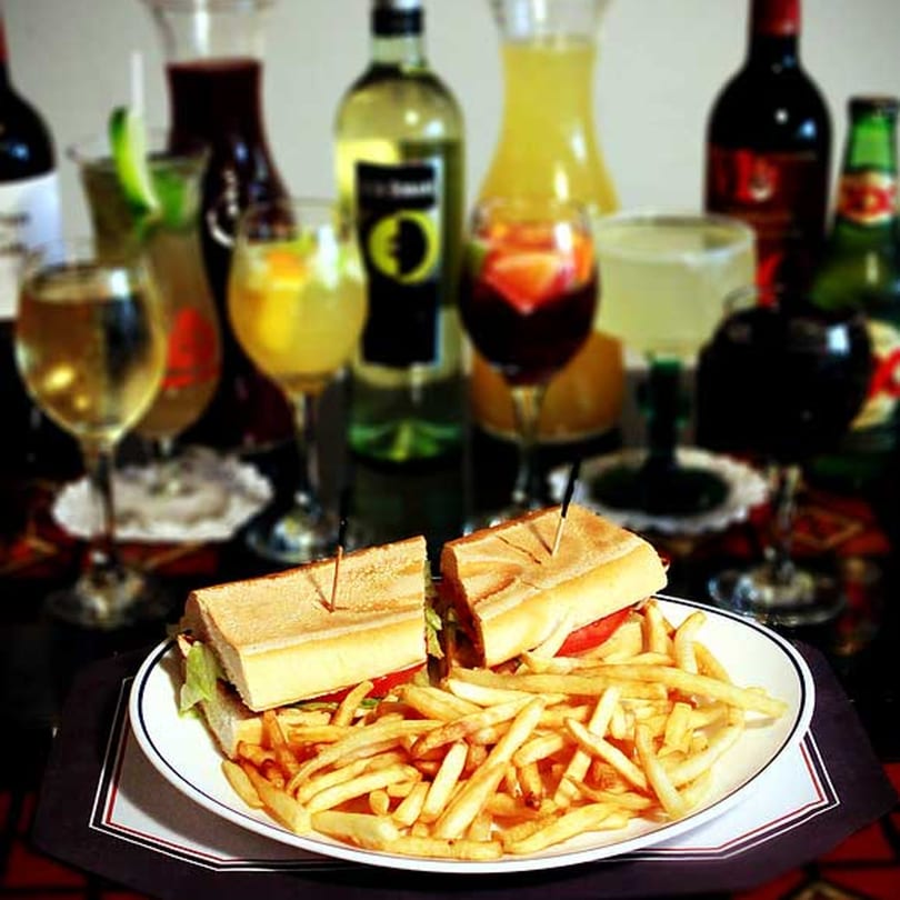A Cuban sandwich with fries on a white plate, surrounded by various drinks and bottles.